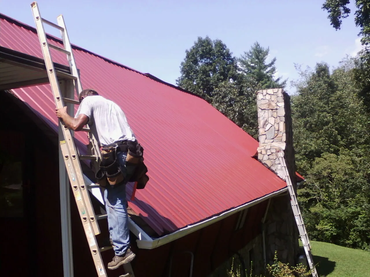 Roofer on a ladder beside a red metal roof and stone chimney — Soffit & Fascia Repair by Roofing Brothers in New London